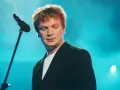 Grzegorz Ciechowski, w czarnym garniturze, pochyla się nad mikrofonem. Jego niezapomniane utwory wciąż brzmią w sercach fanów.