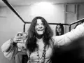 Janis Joplin z kieliszkiem w dłoni, uśmiechnięta. Jej charakterystyczne włosy i naszyjniki przywodzą na myśl jej niezapomniane utwory.