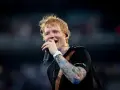 Ed Sheeran śpiewa z mikrofonem, jego ręka ozdobiona jest kolorowymi tatuażami.