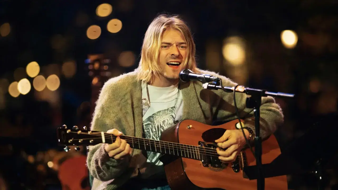 Kurt Cobain śpiewa i gra na gitarze akustycznej, ubrany w sweter i t-shirt.