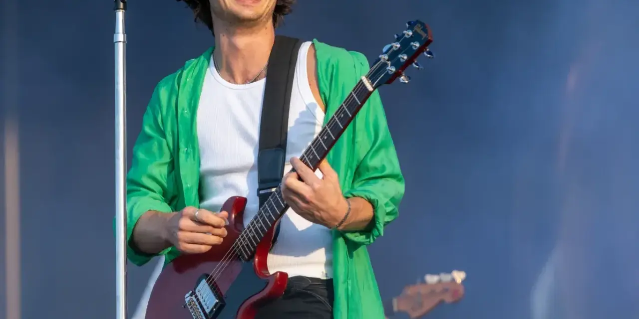 Joe Keery, w zielonej koszuli i białej podkoszulce, gra na czerwonej gitarze elektrycznej na scenie.