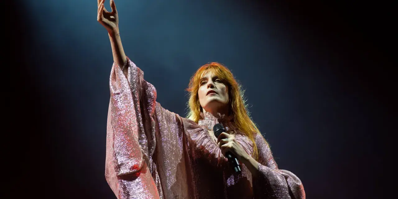 Florence Welch z Florence and the Machine na koncercie, w błyszczącej sukni, z uniesioną ręką.