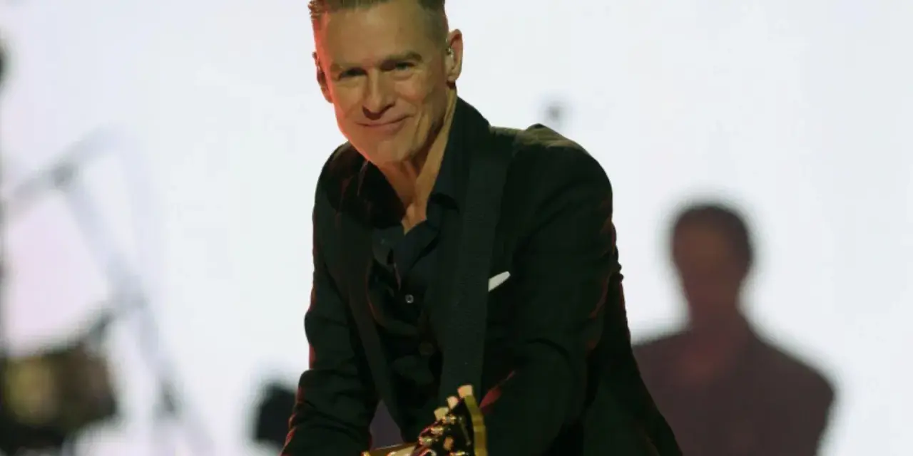 Bryan Adams w czarnym garniturze gra na gitarze. Sprawdź jego nadchodzące wydarzenia i koncerty.