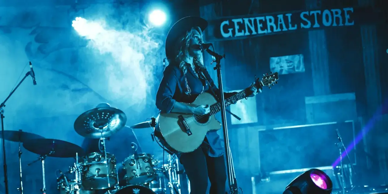 Artystka w kapeluszu gra na gitarze, wykonując najpopularniejsze piosenki country. W tle perkusja i napis "GENERAL STORE".