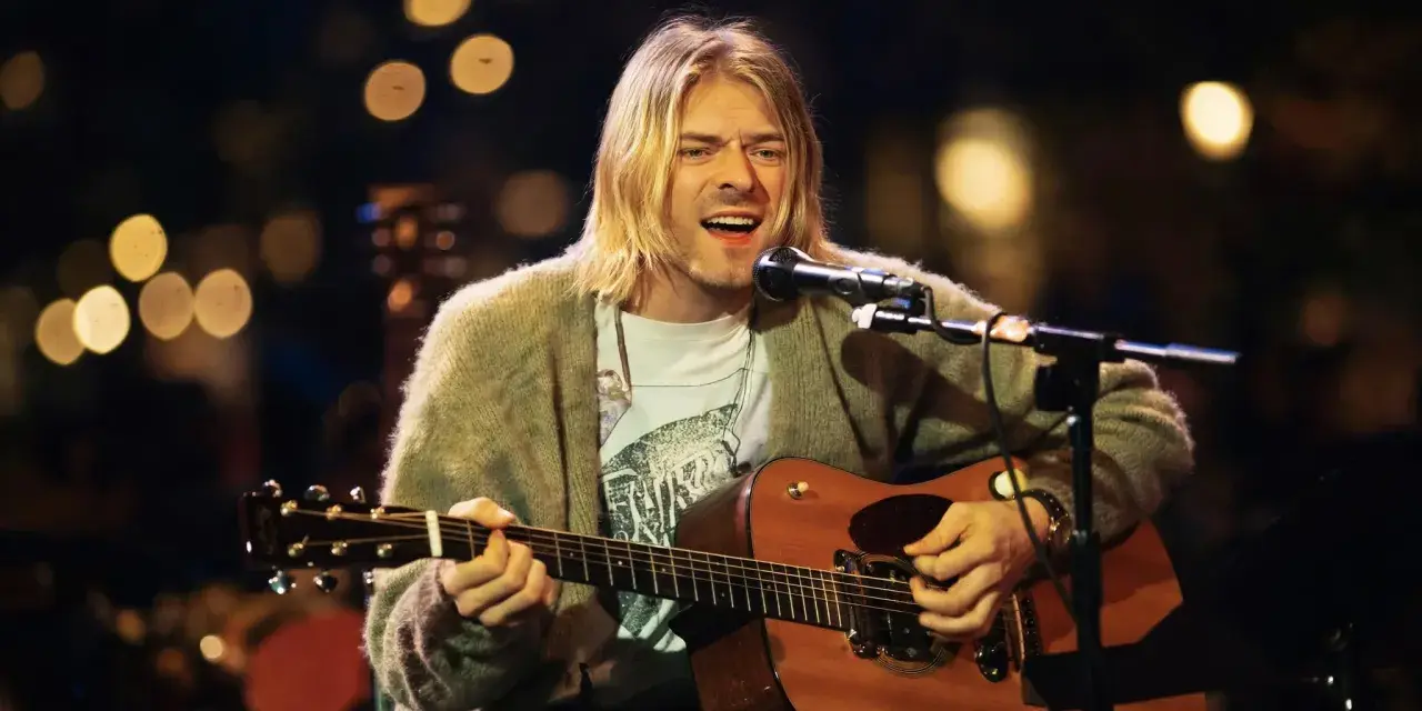 Kurt Cobain śpiewa i gra na gitarze akustycznej, ubrany w sweter i t-shirt.
