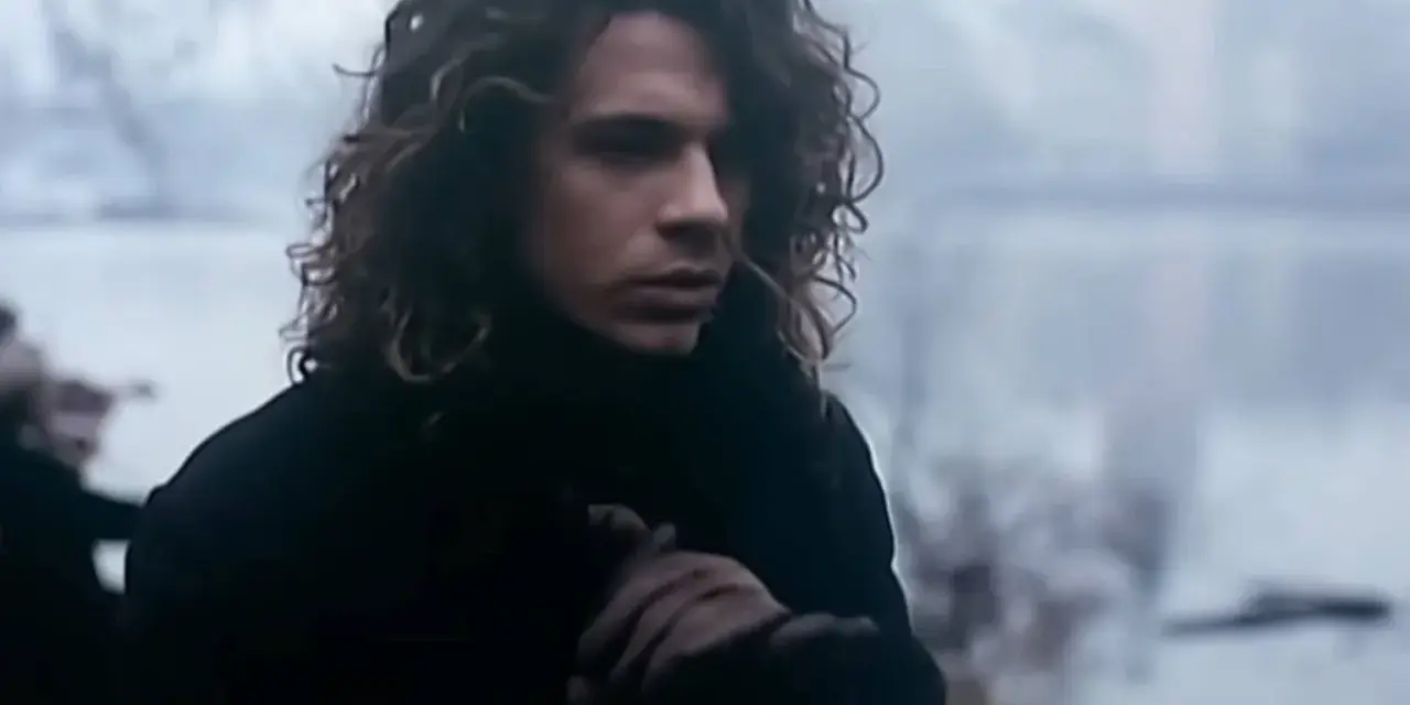 Mężczyzna z kręconymi włosami w czarnym płaszczu, trzymający się za rękę, jakby chciał powiedzieć "never tear us apart".