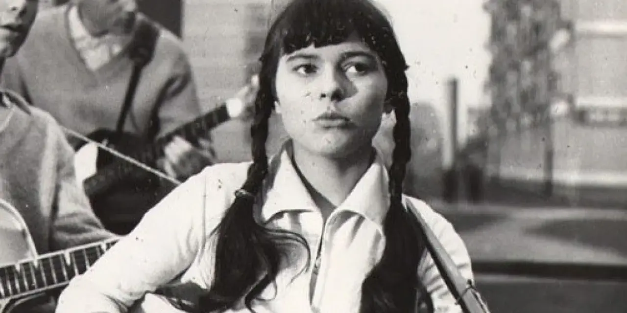 Młoda Karin Stanek z warkoczami śpiewa, grając na gitarze. W tle zespół muzyczny.