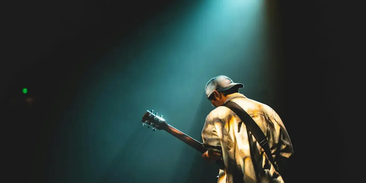 Mężczyzna w czapce z daszkiem gra na gitarze pod światłem reflektora, tworząc atmosferę jak z najsłynniejszych utworów rockowych.