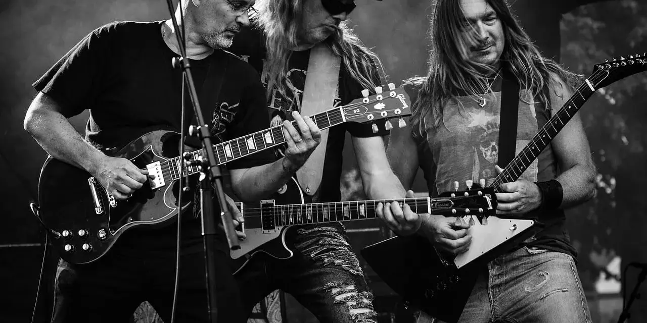 Trzech gitarzystów grających na scenie, przypominających klimatem ballady rockowe lat 80.