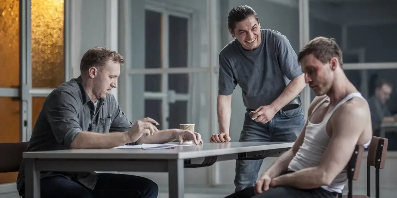 Dario ślepnie od świateł, siedząc przy stole z dokumentami, podczas gdy drugi mężczyzna śmieje się, a trzeci, z podbitym okiem, patrzy w dół.