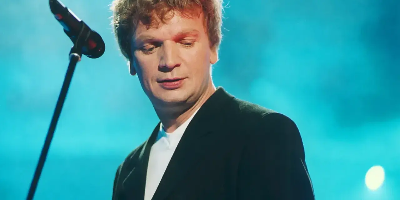 Grzegorz Ciechowski, w czarnym garniturze, pochyla się nad mikrofonem. Jego niezapomniane utwory wciąż brzmią w sercach fanów.
