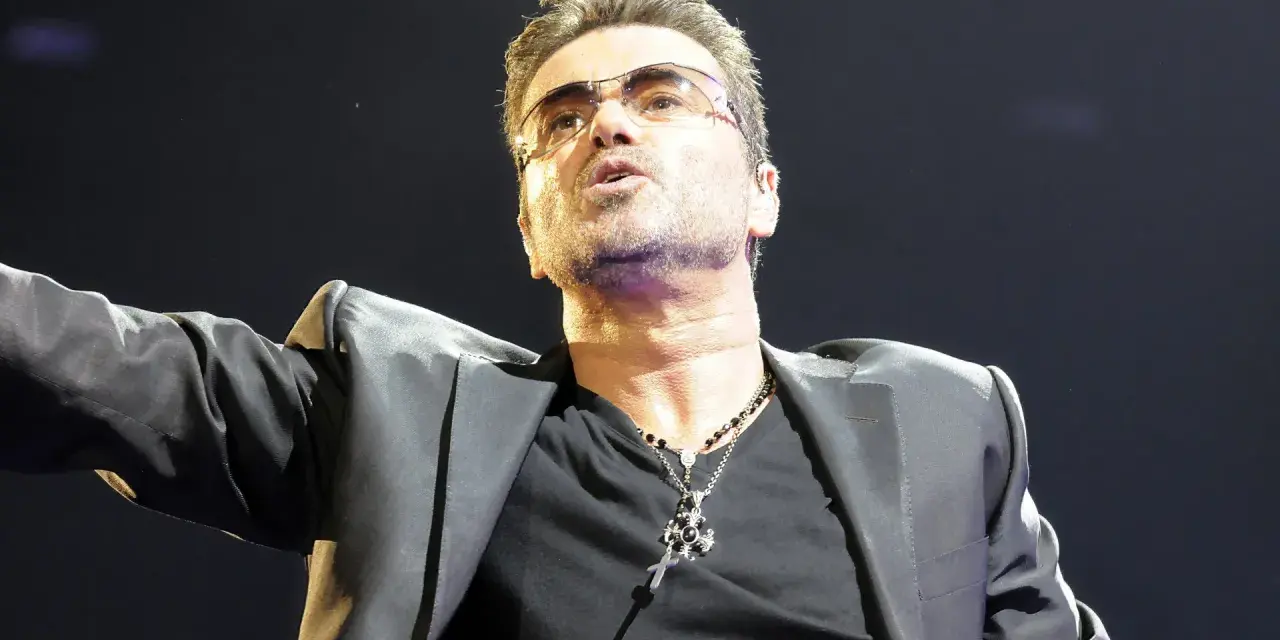 George Michael w okularach, w czarnej marynarce i z naszyjnikiem, śpiewający swoje niezapomniane utwory.