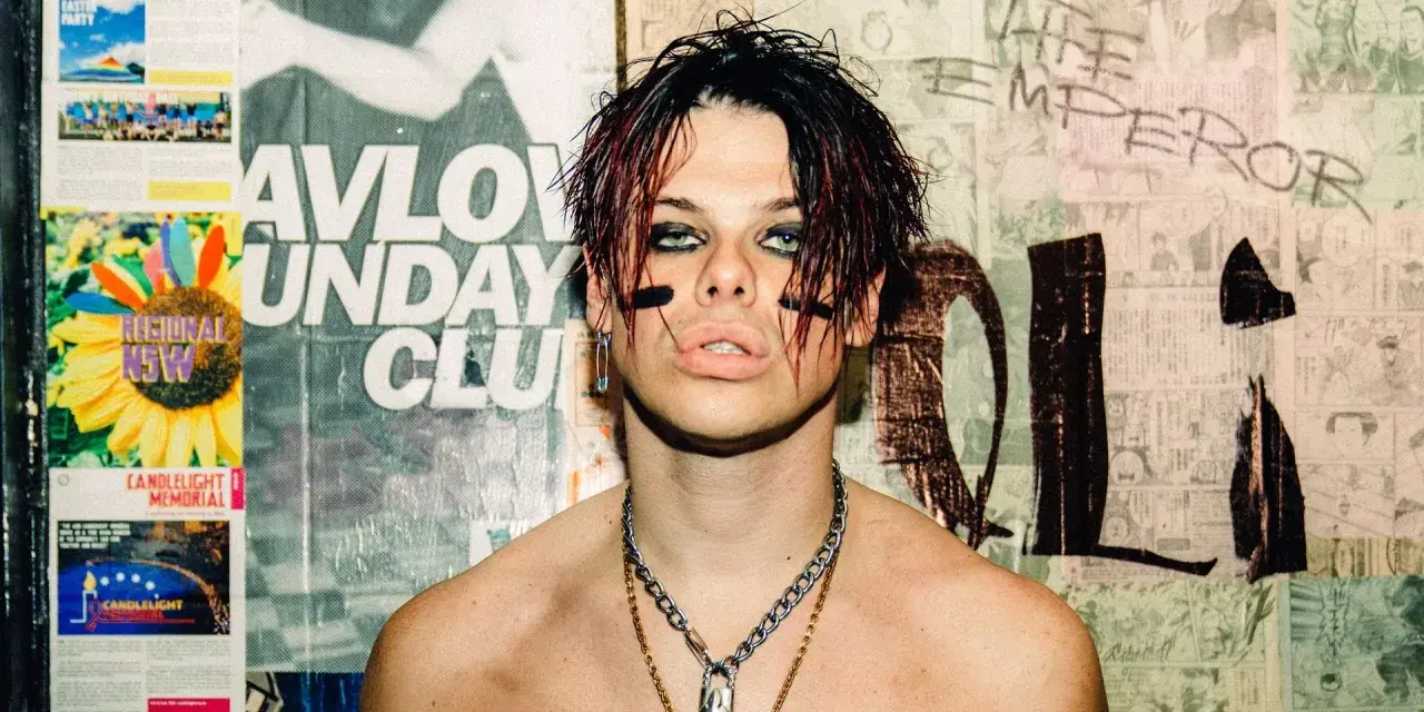 Yungblud, "The Emperor", z charakterystycznym makijażem i łańcuchami, na tle plakatów.