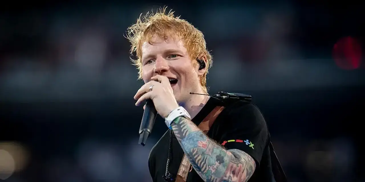 Ed Sheeran śpiewa z mikrofonem, jego ręka ozdobiona jest kolorowymi tatuażami.
