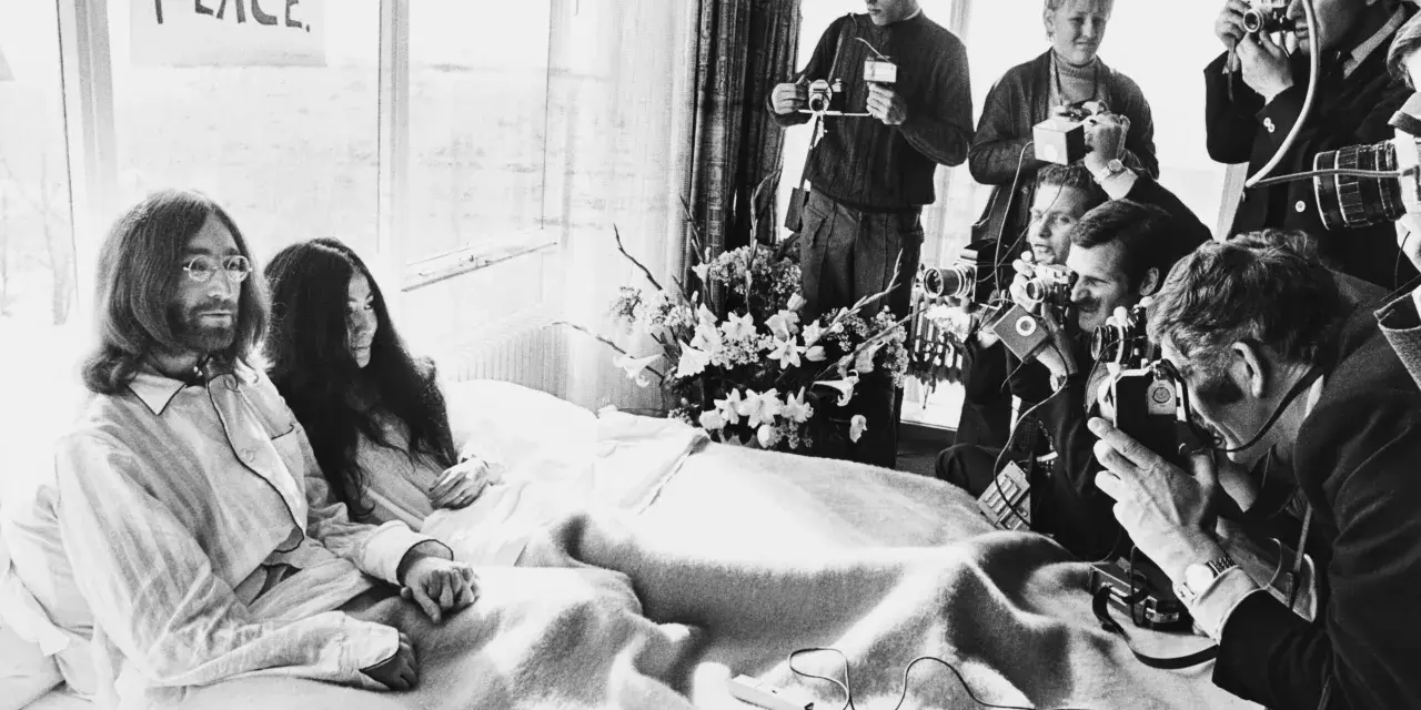 John Lennon i Yoko Ono w łóżku, otoczeni przez reporterów z aparatami. Na ścianie wisi plakat "BED PEACE".