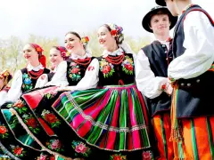 Uśmiechnięci młodzi ludzie w barwnych strojach ludowych, prezentujący polskie tańce narodowe.
