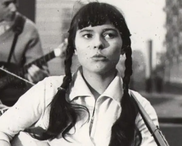 Młoda Karin Stanek z warkoczami śpiewa, grając na gitarze. W tle zespół muzyczny.