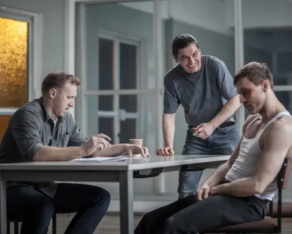 Dario ślepnie od świateł, siedząc przy stole z dokumentami, podczas gdy drugi mężczyzna śmieje się, a trzeci, z podbitym okiem, patrzy w dół.