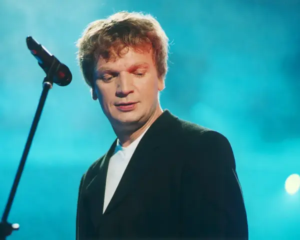 Grzegorz Ciechowski, w czarnym garniturze, pochyla się nad mikrofonem. Jego niezapomniane utwory wciąż brzmią w sercach fanów.