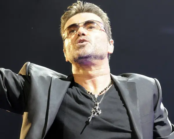 George Michael w okularach, w czarnej marynarce i z naszyjnikiem, śpiewający swoje niezapomniane utwory.