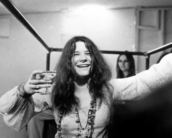 Janis Joplin z kieliszkiem w dłoni, uśmiechnięta. Jej charakterystyczne włosy i naszyjniki przywodzą na myśl jej niezapomniane utwory.