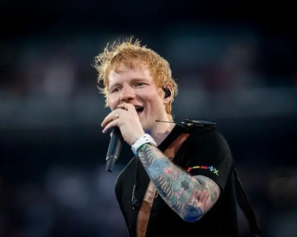 Ed Sheeran śpiewa z mikrofonem, jego ręka ozdobiona jest kolorowymi tatuażami.