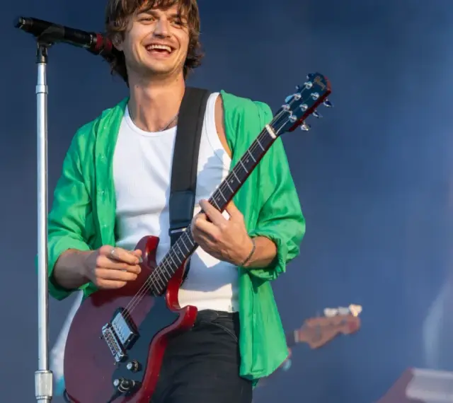 Joe Keery, w zielonej koszuli i białej podkoszulce, gra na czerwonej gitarze elektrycznej na scenie.