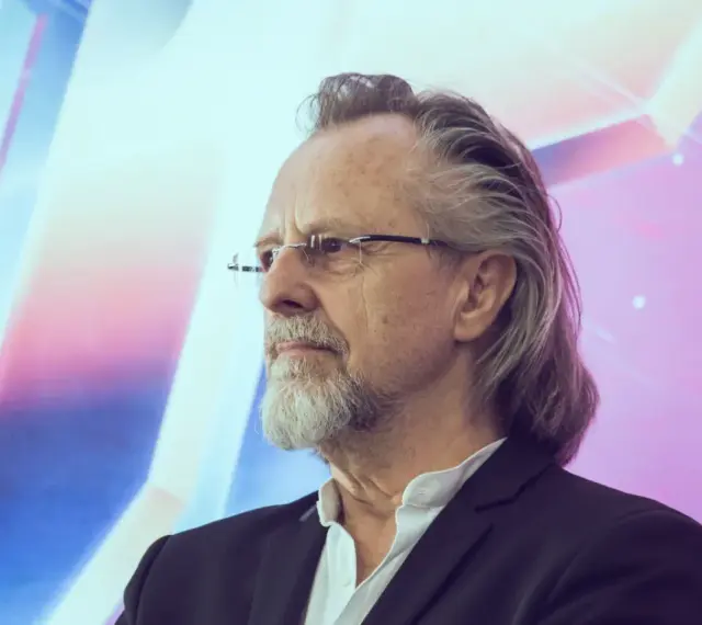 Portret mężczyzny z siwą brodą i długimi włosami, jednego z wybitnych kompozytorów muzyki filmowej, w okularach i czarnym garniturze.