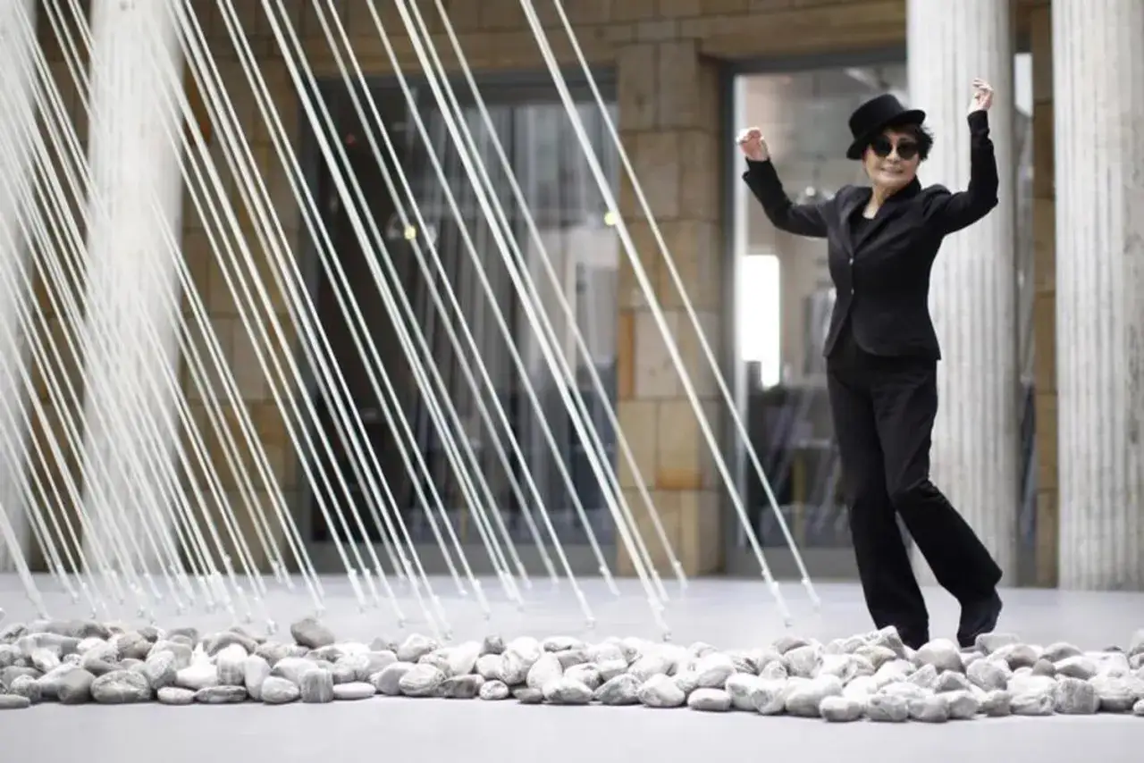 Yoko Ono, w czarnym garniturze i kapeluszu, tańczy wśród białych sznurków i kamieni, przypominając o duchu Johna Lennona.
