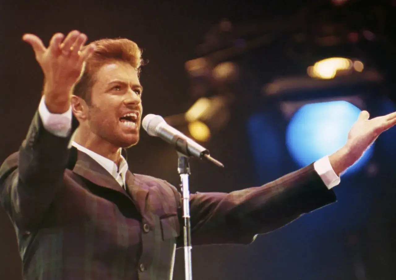 George Michael śpiewa, wykonując swoje niezapomniane utwory. Jego dłonie unoszą się w geście, a głos niesie się przez mikrofon.