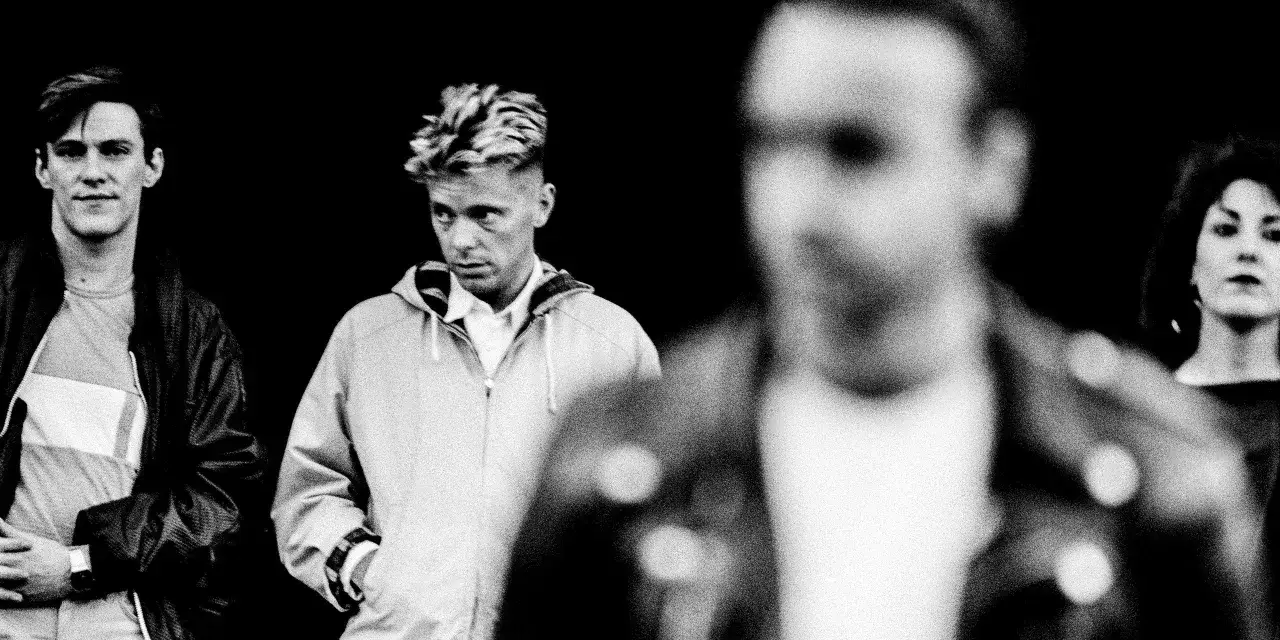 Cztery postacie w stylu lat 80. na tle czerni. Jeden z nich, z charakterystyczną fryzurą, przypomina wokalistę New Order w dzień 