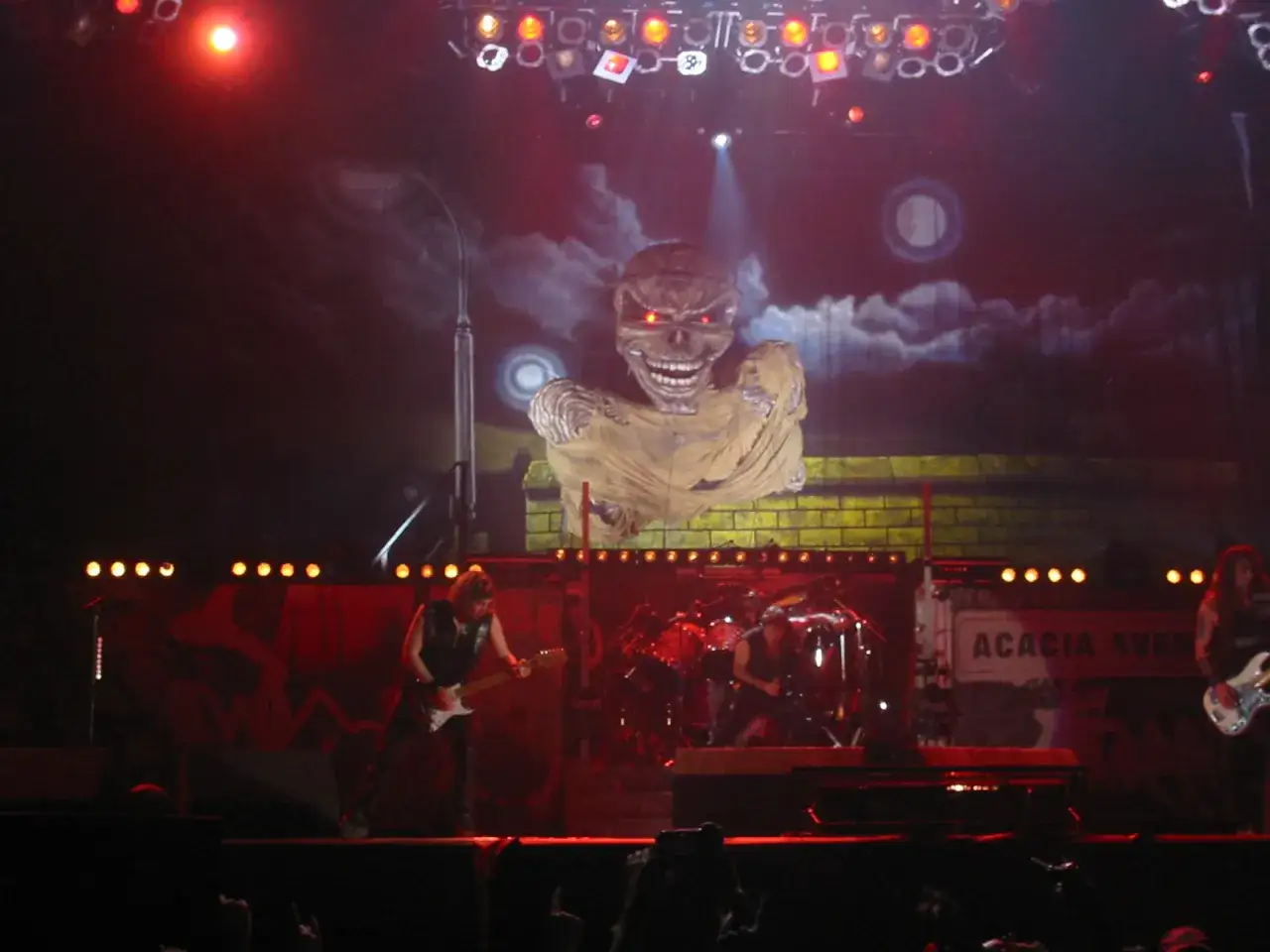 Koncert Iron Maiden. Na scenie Eddie, gitarzysta i perkusista. W tle wielki Eddie z płonącymi oczami.