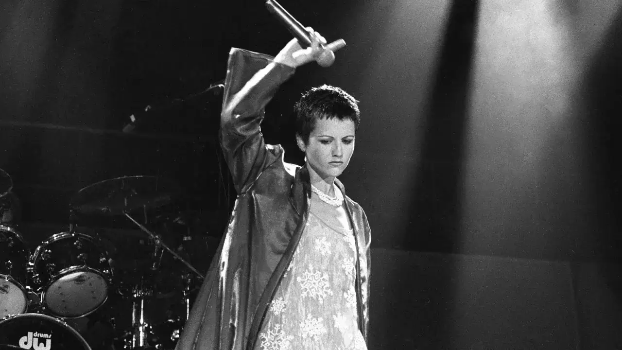 Dolores O'Riordan z The Cranberries, z mikrofonem w dłoni, na tle perkusji. Jej charakterystyczne utwory brzmią w uszach.