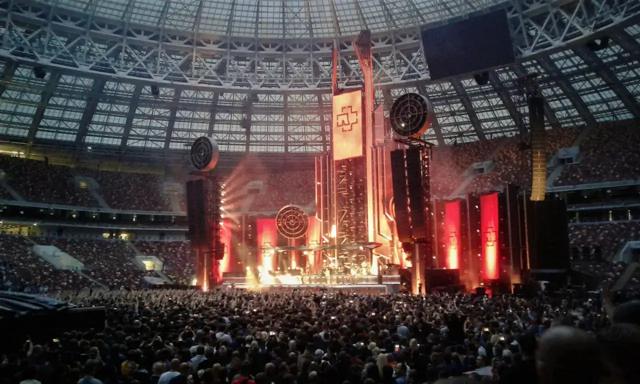 Koncert Rammstein - Deutschland. Ogromny tłum na stadionie, scena z pirotechniką i charakterystycznym logo zespołu.
