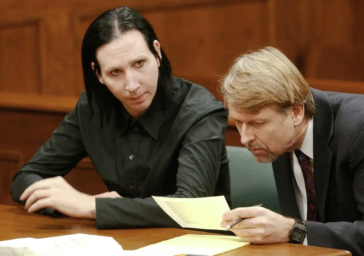 Marilyn Manson bez makijażu, z czarnymi włosami i w czarnej koszuli, siedzi przy stole z prawnikiem przeglądającym dokumenty.