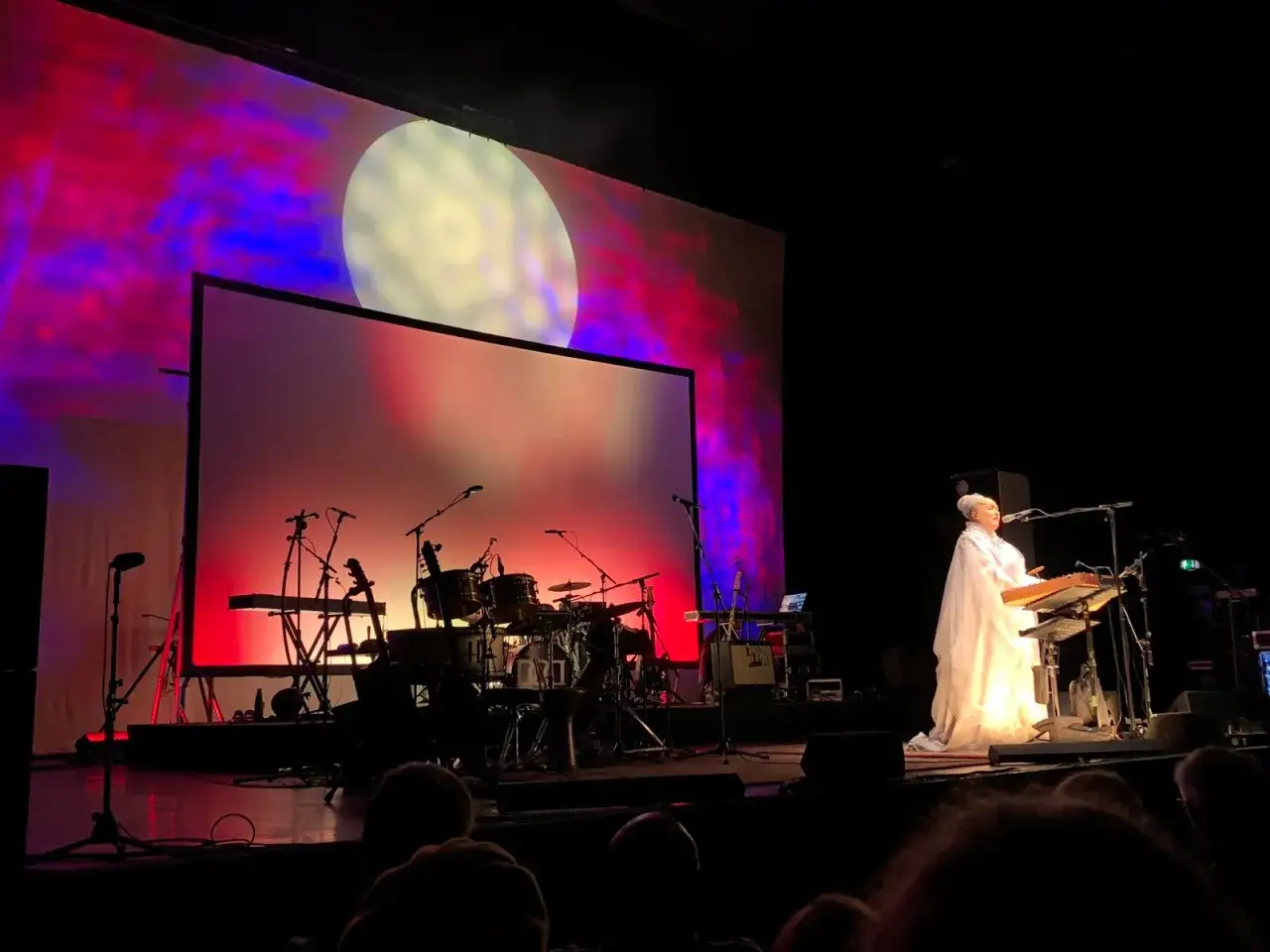 Lisa Gerrard w białej kreacji gra na keyboardzie podczas koncertu Dead Can Dance. Tło sceny rozświetla projekcja z wielką, żółtą tarczą.