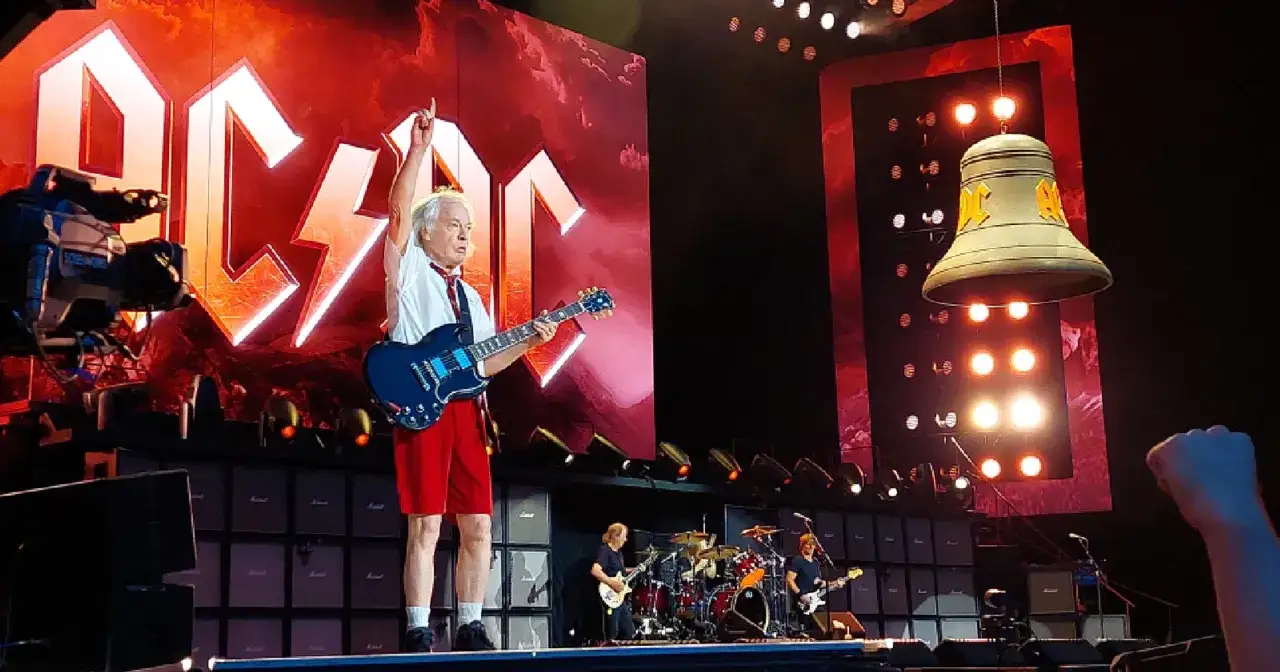 Angus Young z AC/DC gra na gitarze podczas koncertu w Polsce. Na scenie wielki dzwon i logo zespołu.