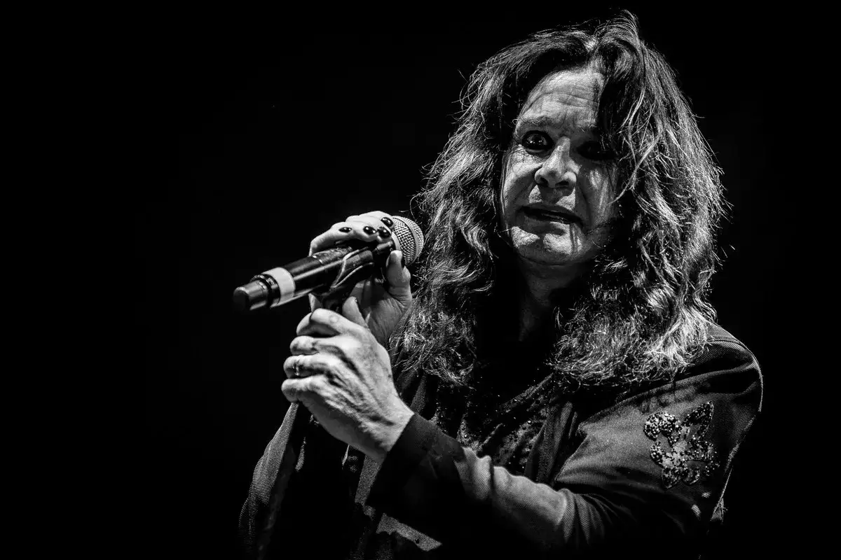Ozzy Osbourne śpiewa do mikrofonu, jego charakterystyczna fryzura i makijaż podkreślają jego sceniczny wizerunek.