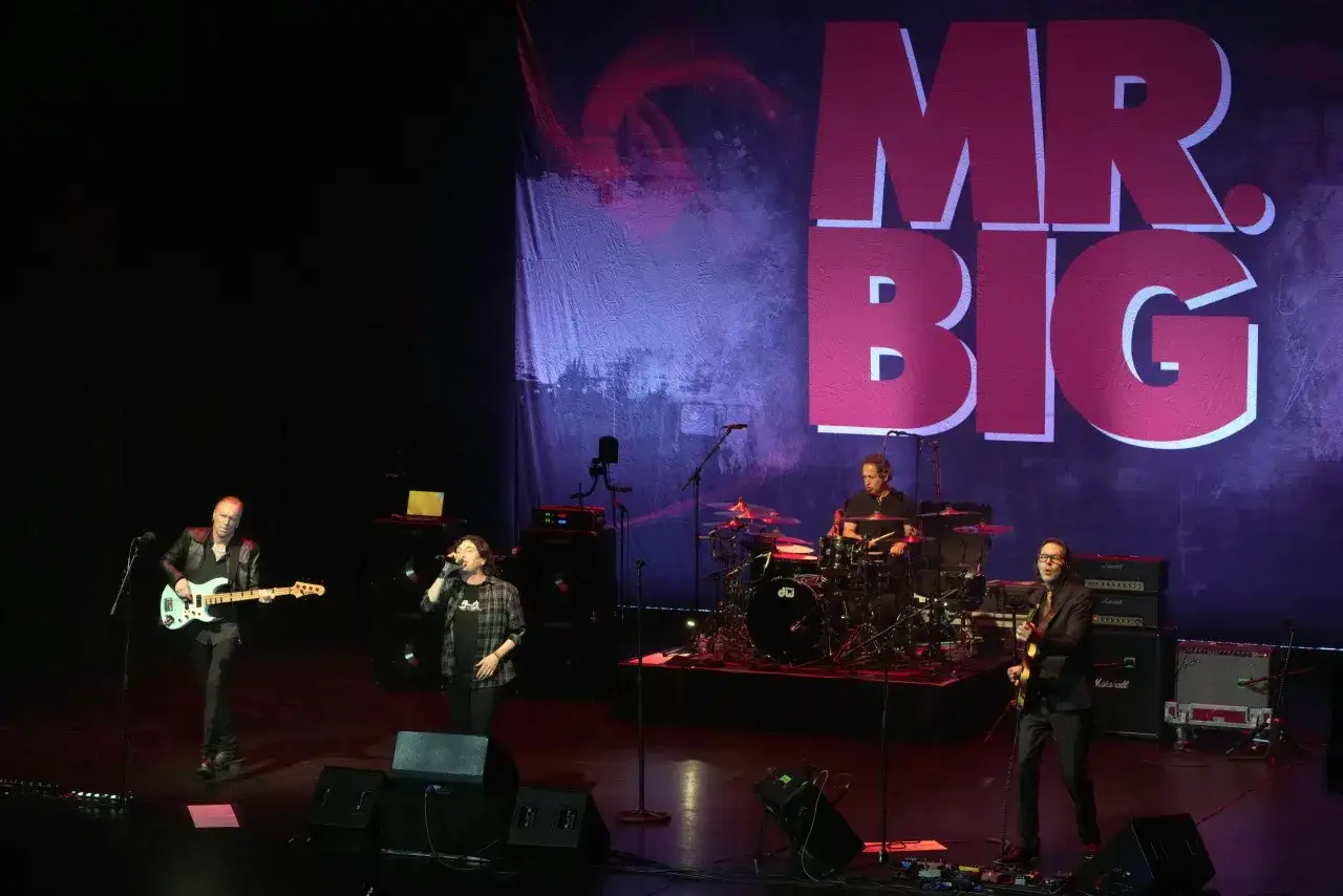 Zespół Mr. Big na scenie, grający dla fanów. W tle wielki napis 