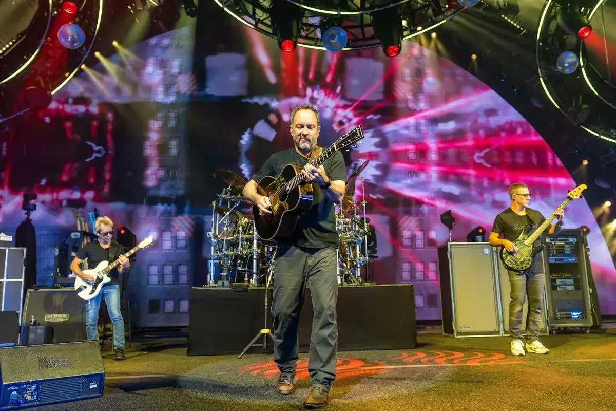 Dave Matthews Band (DMB) na scenie, grający na gitarach i basie. Koncertowa atmosfera z efektami świetlnymi.