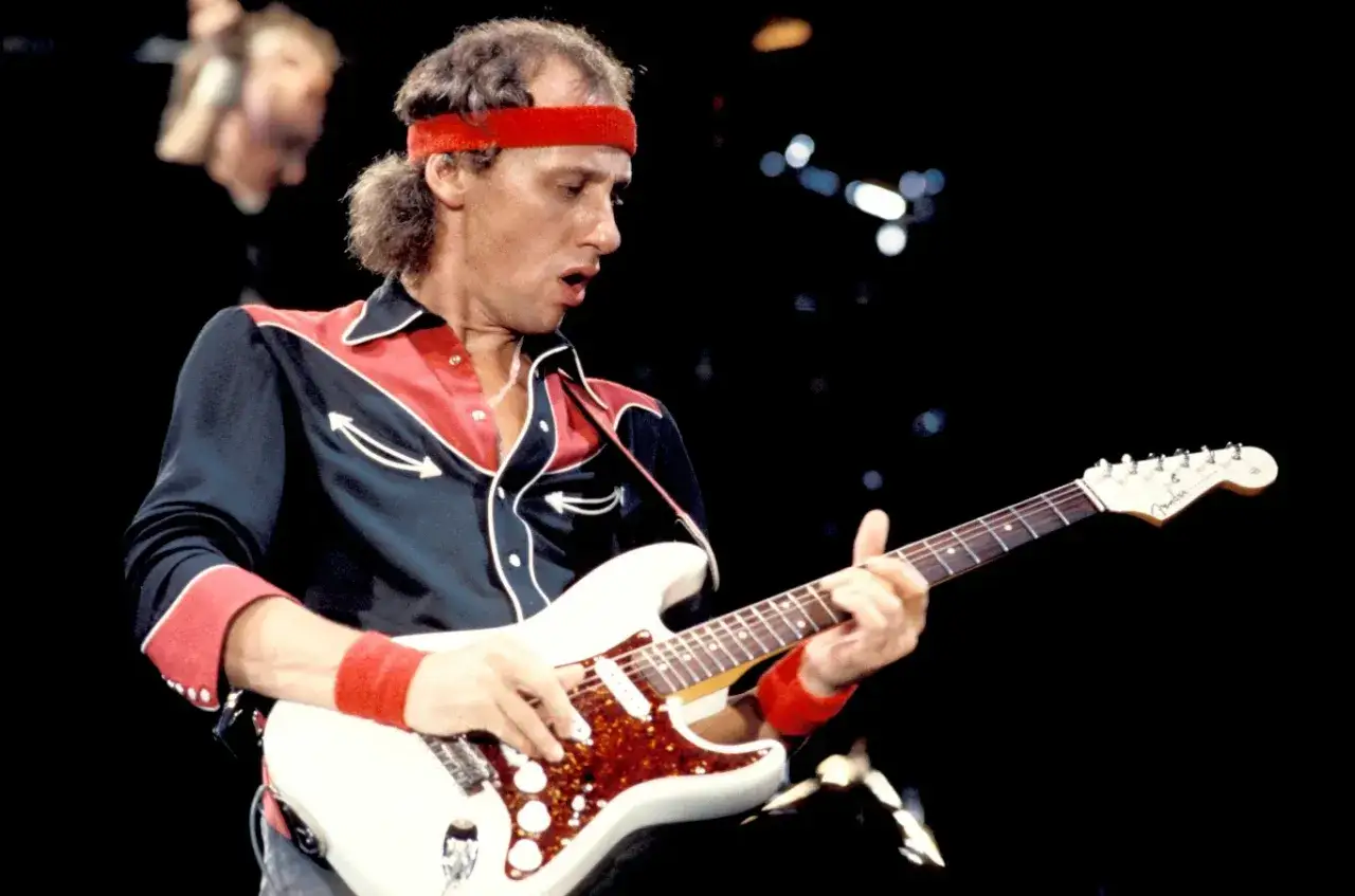 Mark Knopfler w czerwonej opasce na głowie i charakterystycznej koszuli gra na białej gitarze. Jego utwory to klasyka rocka.