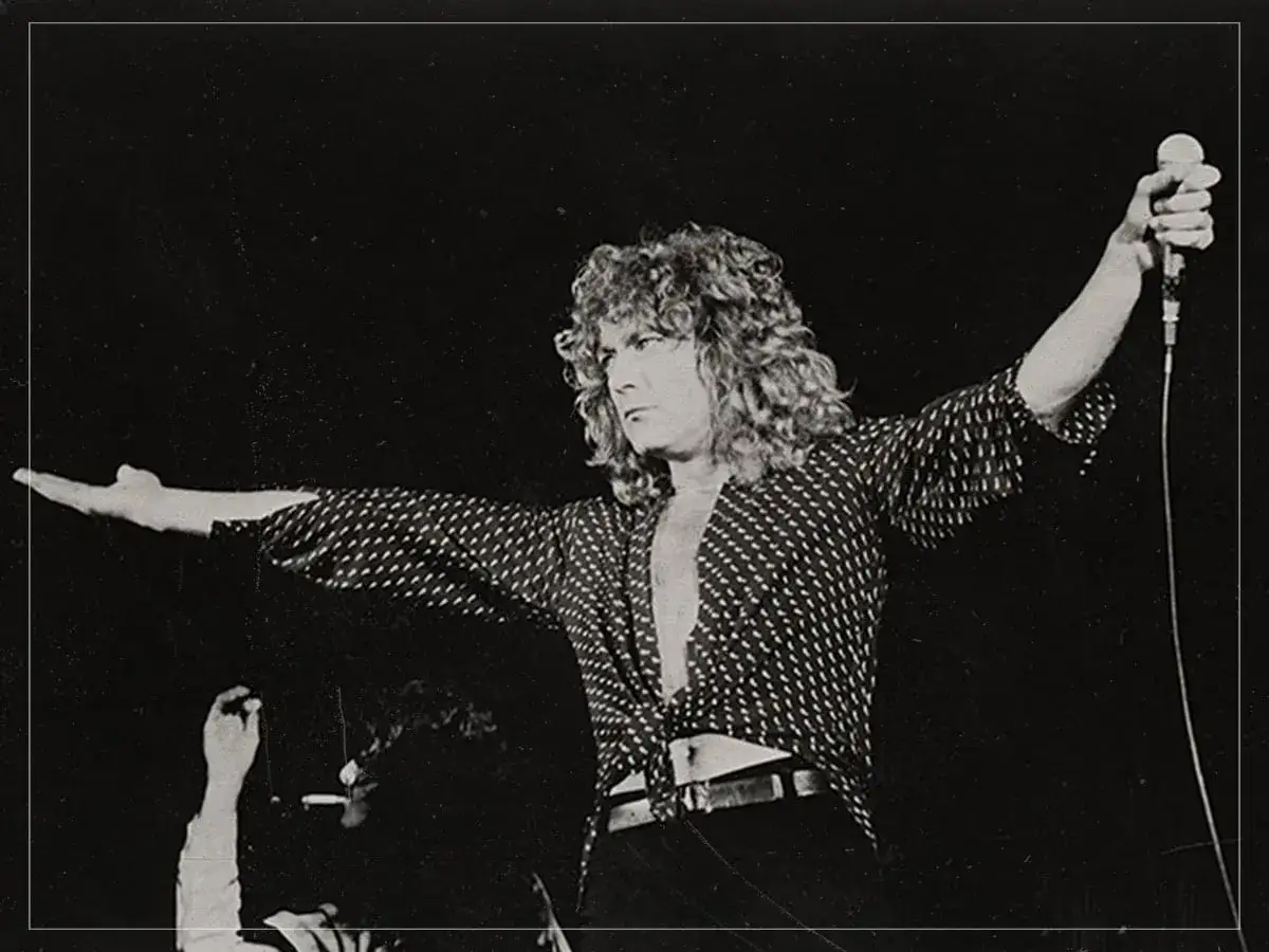 Wokalista Led Zeppelin, Robert Plant, z rozwianymi włosami i w charakterystycznej koszuli, śpiewa do mikrofonu na koncercie.