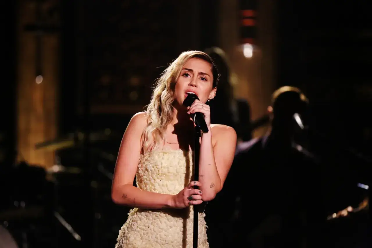 Miley Cyrus w kremowej sukni wykonuje fajne covery znanych piosenek na scenie, z zespołem w tle.