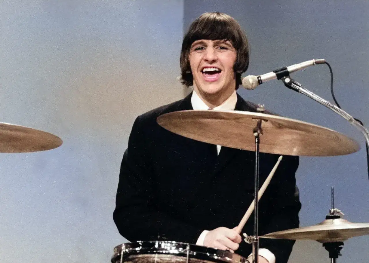 Ringo Starr z The Beatles, w czarnym garniturze, gra na perkusji, śmiejąc się do mikrofonu.