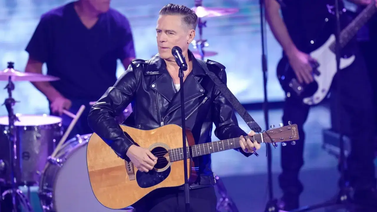 Bryan Adams w skórzanej kurtce gra na gitarze akustycznej, zapowiadając nadchodzące wydarzenia. Na scenie towarzyszy mu zespół.