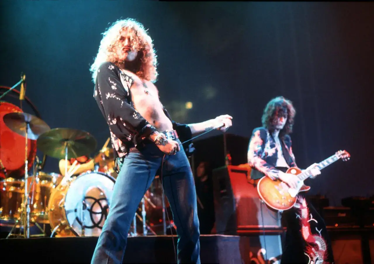 Wokalista Led Zeppelin, Robert Plant, z charakterystyczną burzą blond włosów, śpiewa na scenie. Obok niego Jimmy Page gra na gitarze.